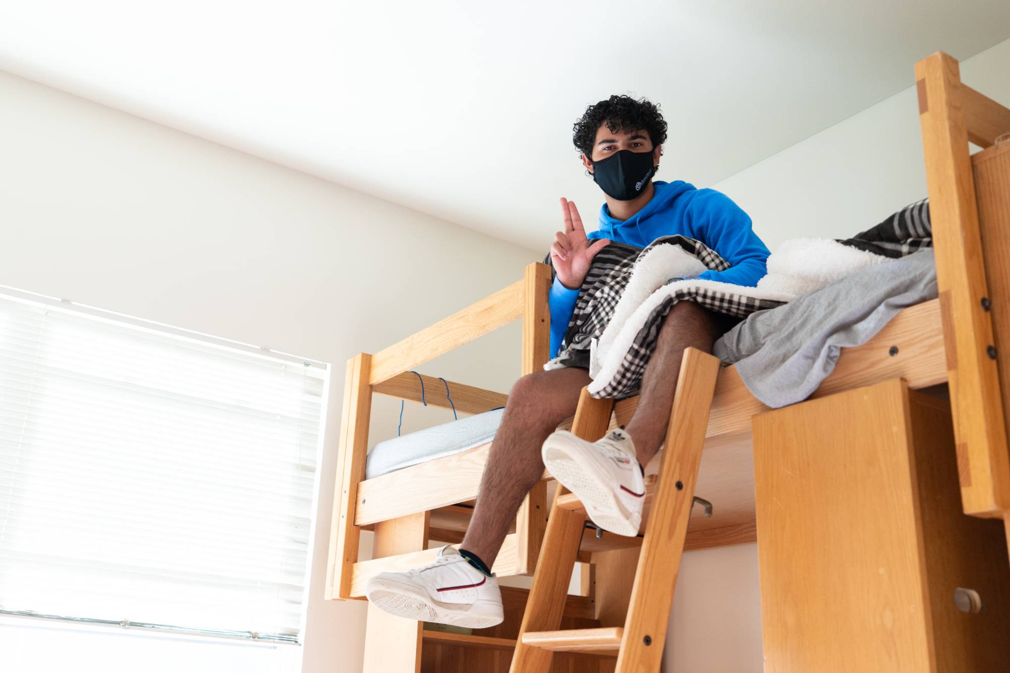 A student sits on his lofted bed in his dorm room, giving a GVSU Anchor Up hand to the camera.